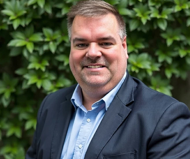 Kenny Schultz in dark blazer smiling outdoors in front of green foliage
