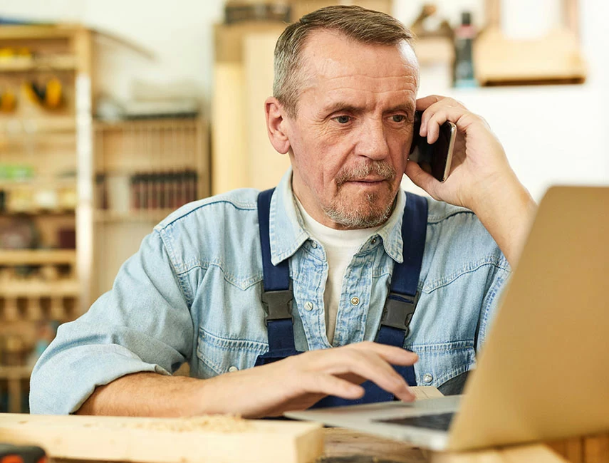 Small business owner in work overalls talking on phone while using laptop in workshop